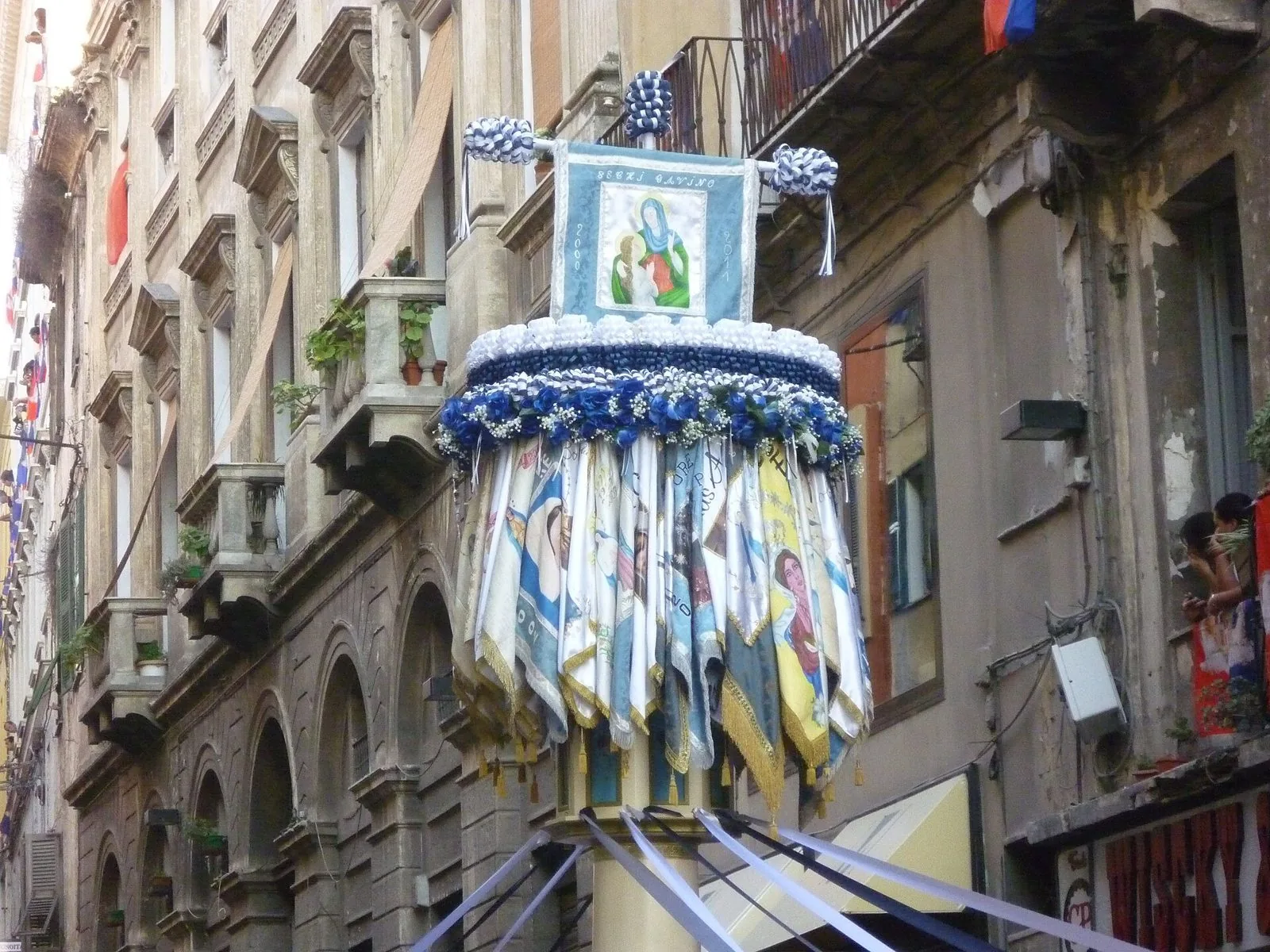 La sommità di un candeliere di Sassari con stendardo della Madonna, corona di fiori blu e bianchi e bandierine colorate, fra i palazzi storici del centro