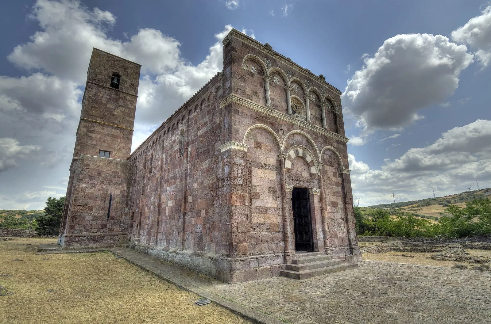 La basilica romanica di Nostra Signora di Tergu, con facciata ad archetti in pietra rossa e bianca e campanile squadrato, sotto un cielo con nuvole